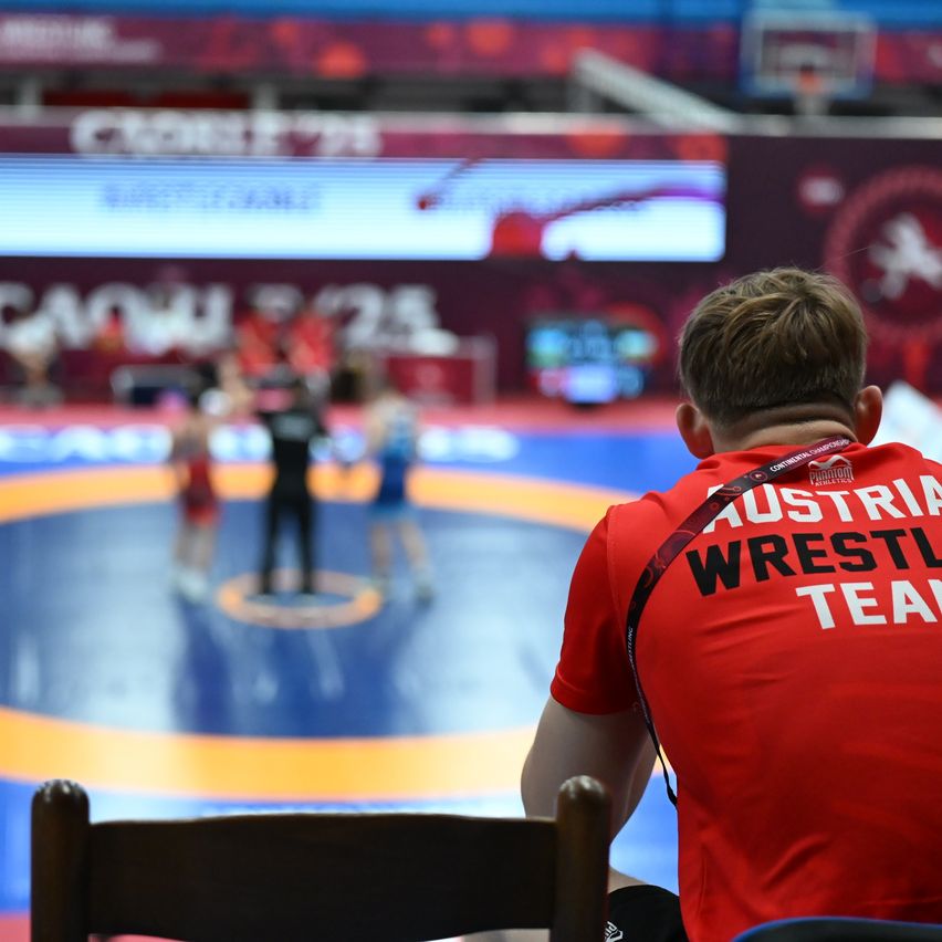 Eine Person in einem roten Trikot der österreichischen Wrestling-Nationalmannschaft beobachtet von der Seitenlinie, wie zwei Konkurrenten auf einem blau-orangefarbenen Ring kämpfen.