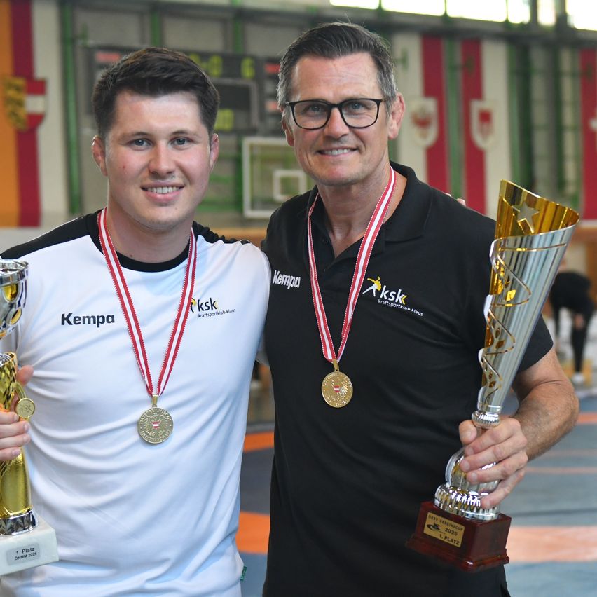 Zwei Männer halten Trophäen in einer Turnhalle und lächeln. Einer trägt ein weißes T-Shirt mit Kempa, der andere ein schwarzes T-Shirt mit aksk. Beide haben Goldmedaillen.