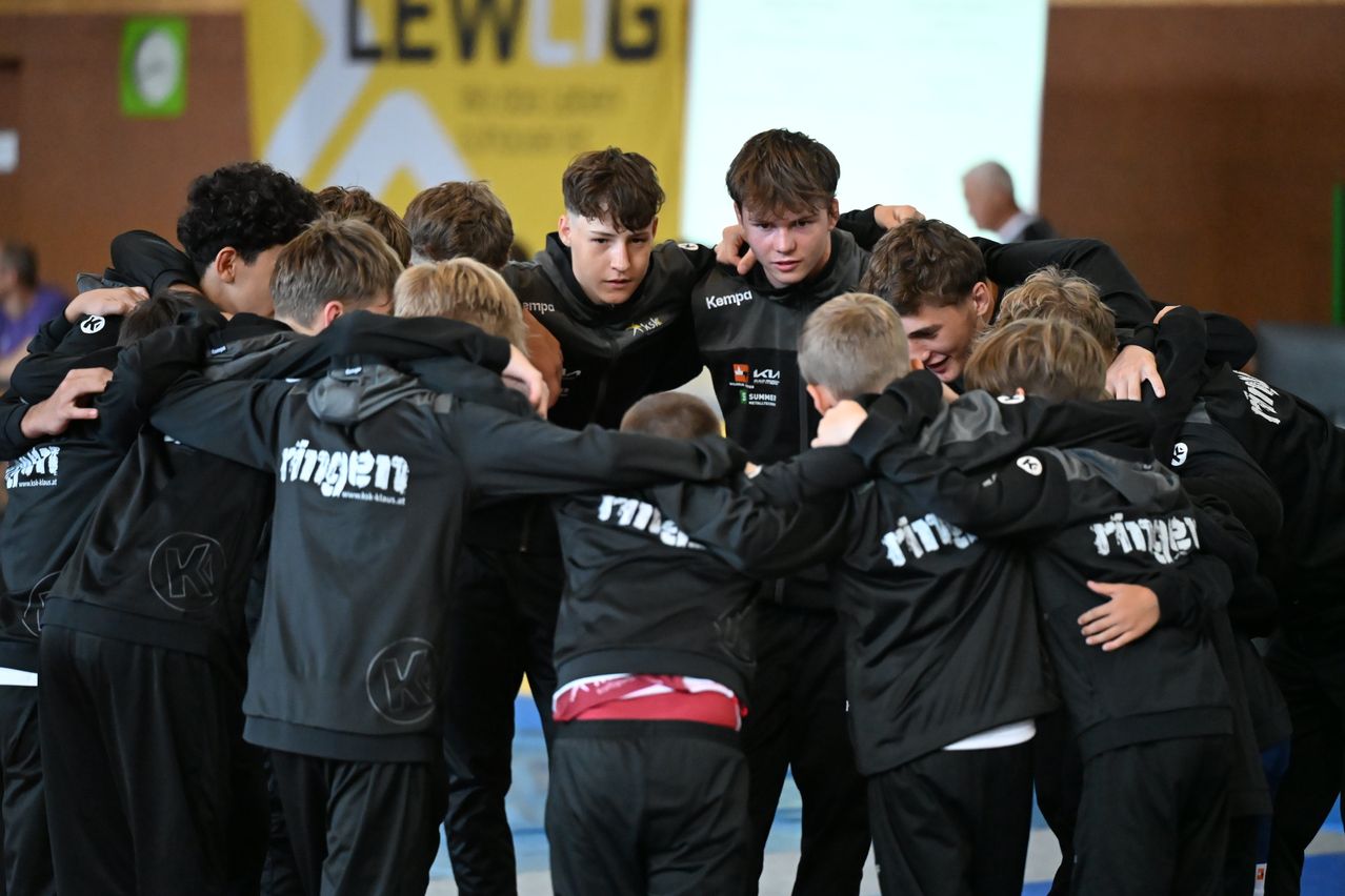 Eine Gruppe junger Athleten in schwarzen Jacken mit verschiedenen Logos steht in einer Turnhalle und bereitet sich auf einen Wettkampf vor.