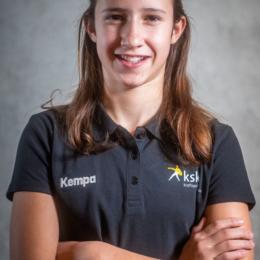 Ein junges Mädchen in einem schwarzen Kempa-Polohemd lächelt mit verschränkten Armen in einem Studio.