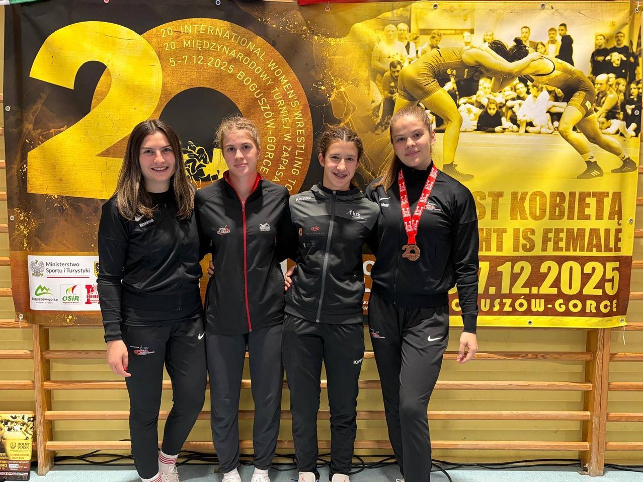 Vier Frauen in schwarzen Jacken und Hosen stehen vor einem großen Banner mit einem Wrestling-Bild. Eine Frau trägt eine Silbermedaille.