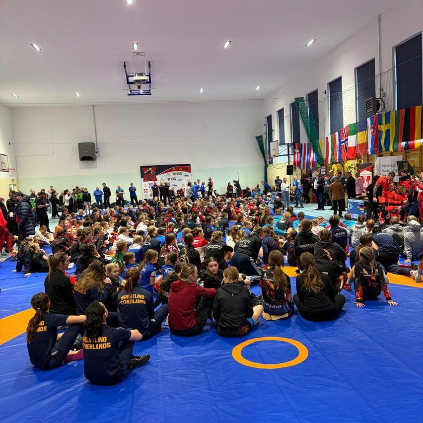 Viele Menschen sind in einer Sporthalle versammelt, einige sitzen auf blauen Matten und andere stehen. Flaggen verschiedener Länder sind an der Wand angebracht. Ein Basketballkorb ist sichtbar.