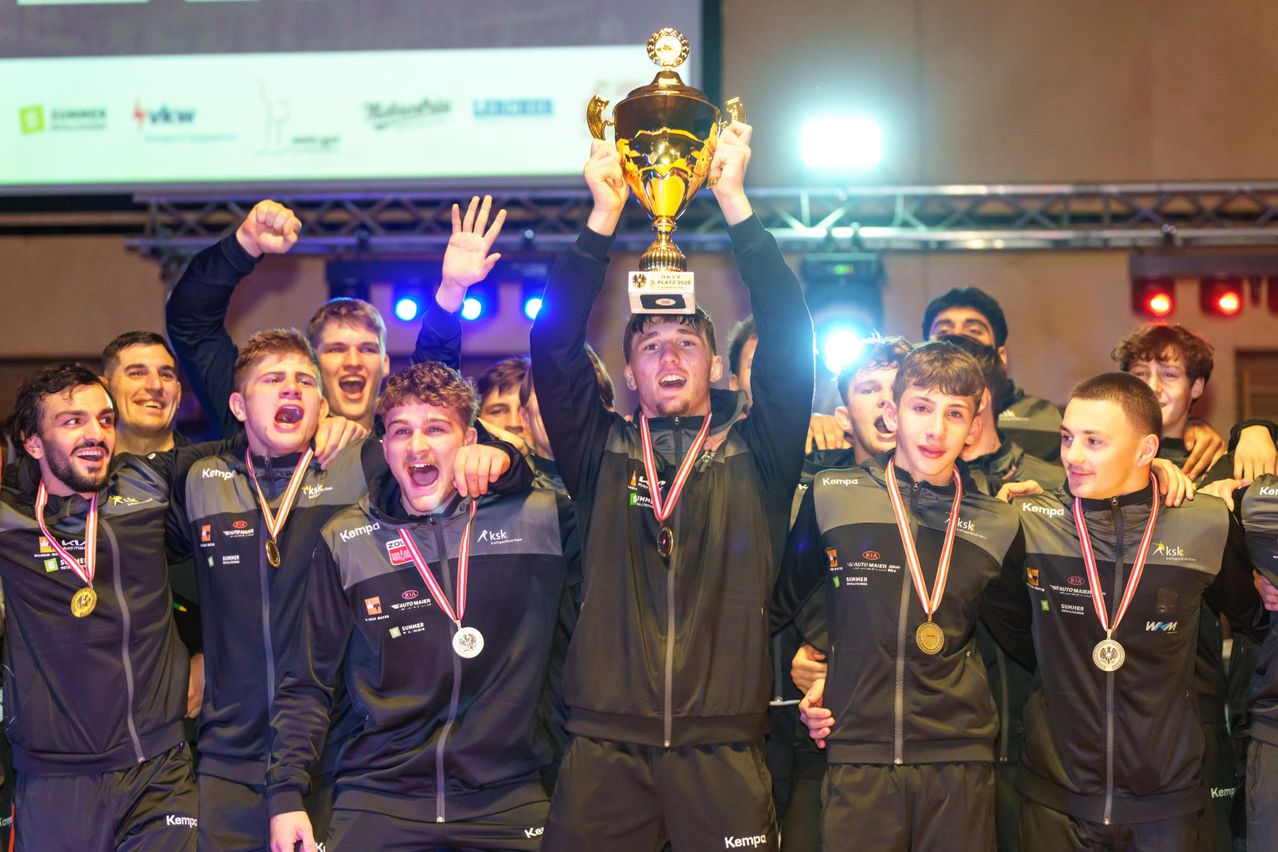 Eine Gruppe junger Männer feiert und hält einen Pokal über ihren Köpfen mit Medaillen um den Hals. Sie tragen schwarze Jacken mit Logos. Der Mann in der Mitte hält den Pokal hoch.