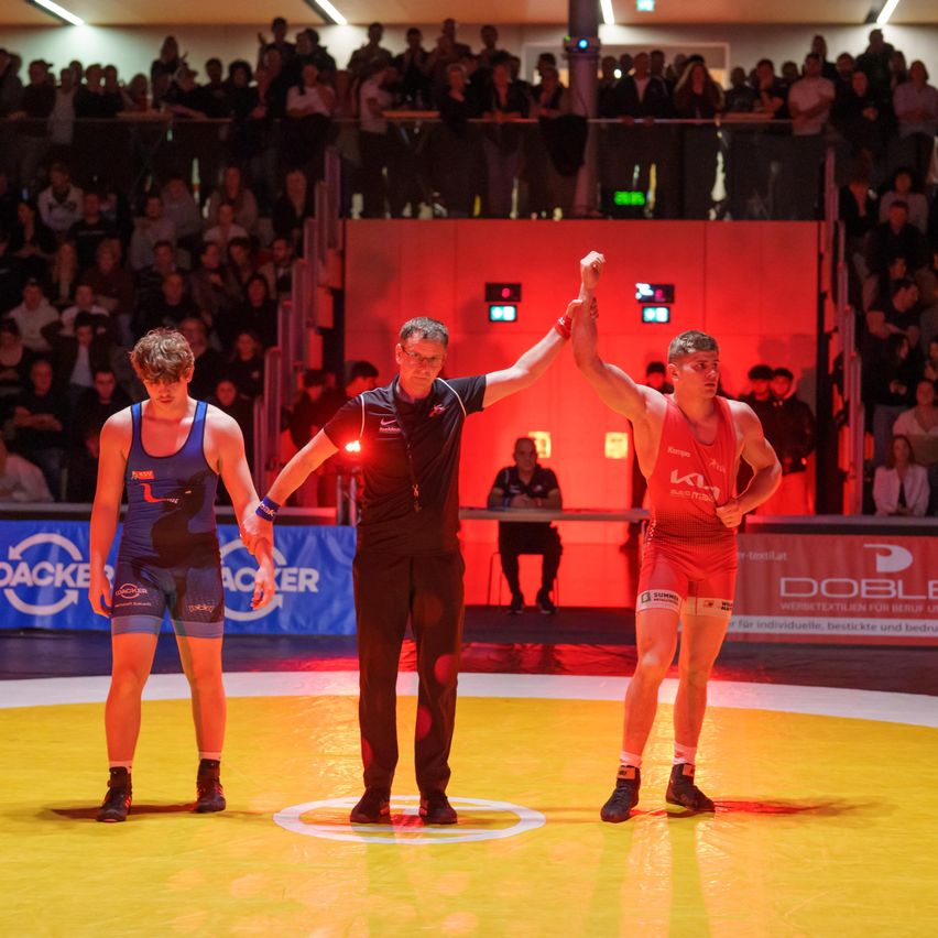 Zwei Männer in roten und blauen Wrestling-Outfits stehen auf einem gelben Teppich mit einem Schiedsrichter. Der Schiedsrichter hebt den Arm eines der Männer, was einen Sieg anzeigt. Ein Publikum schaut von den Tribünen zu. Das Bild hat eine rote und gelbe Beleuchtung.