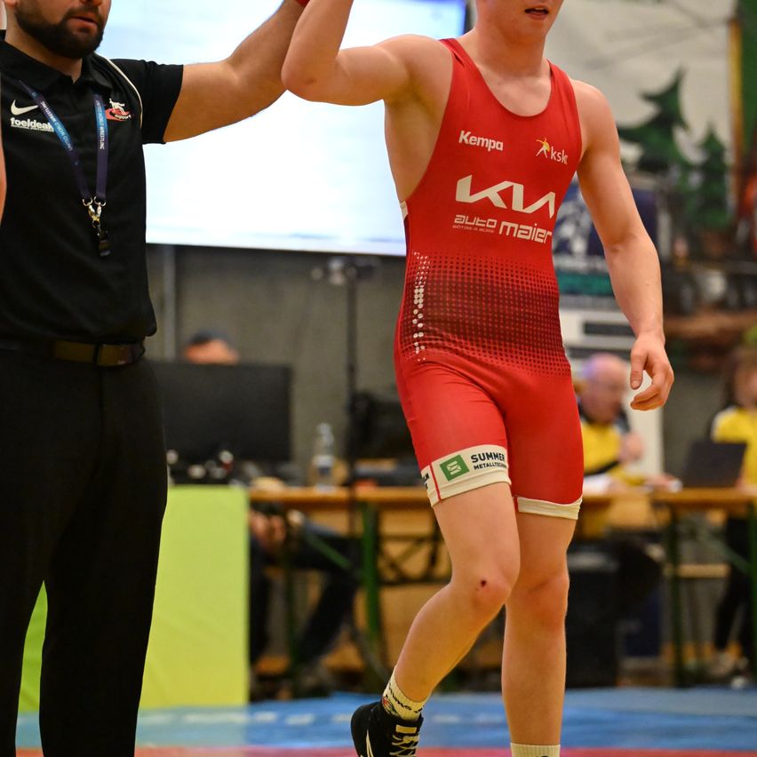 Ein Mann in rotem Wrestling-Equipment steht neben einem Schiedsrichter. Dahinter sitzen Leute an Tischen. Der Raum hat eine große Leinwand, und Bäume sind draußen sichtbar.