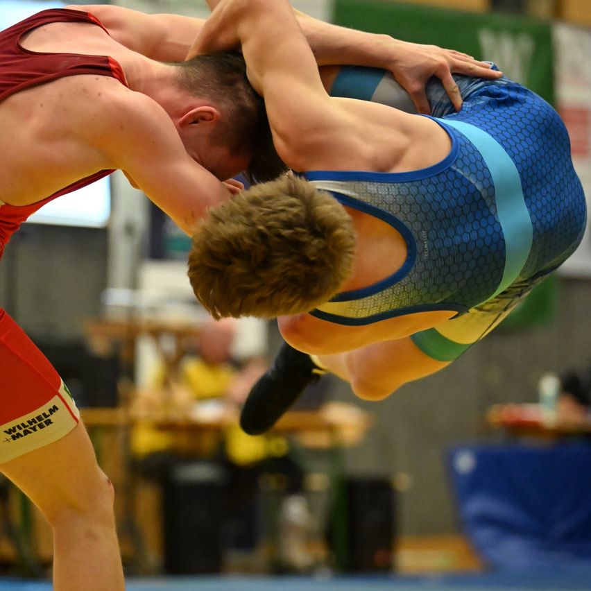 Zwei Ringer kämpfen in einer Indoor-Sporthalle, einer ist in der Luft. Einer trägt ein rotes Shirt und Shorts, der andere ein blaues und graues Trikot. Einige Leute sitzen im Hintergrund und beobachten den Wettkampf.