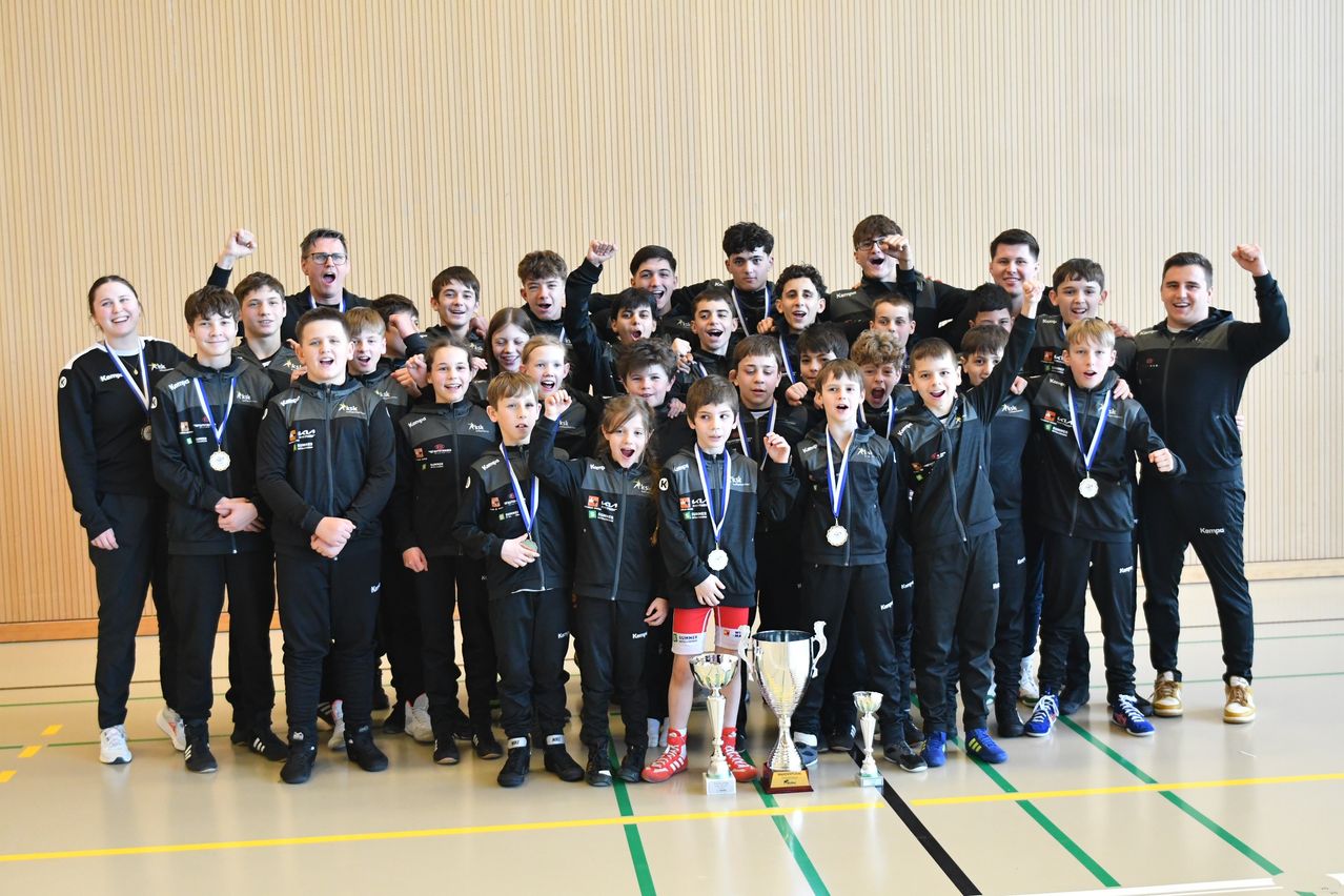 A group of young boys and men pose for a photo in a gym, wearing medals and uniforms, with trophies in front of them.