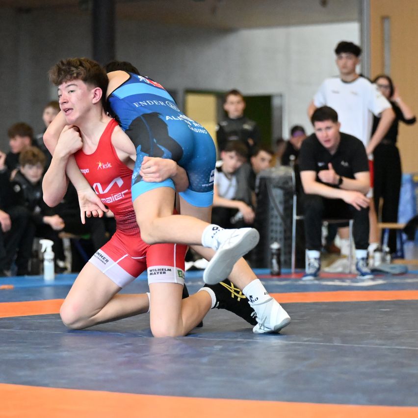 Two young men in red and blue wrestling outfits compete on a blue and orange mat. Spectators watch from the sidelines, some seated and others standing.