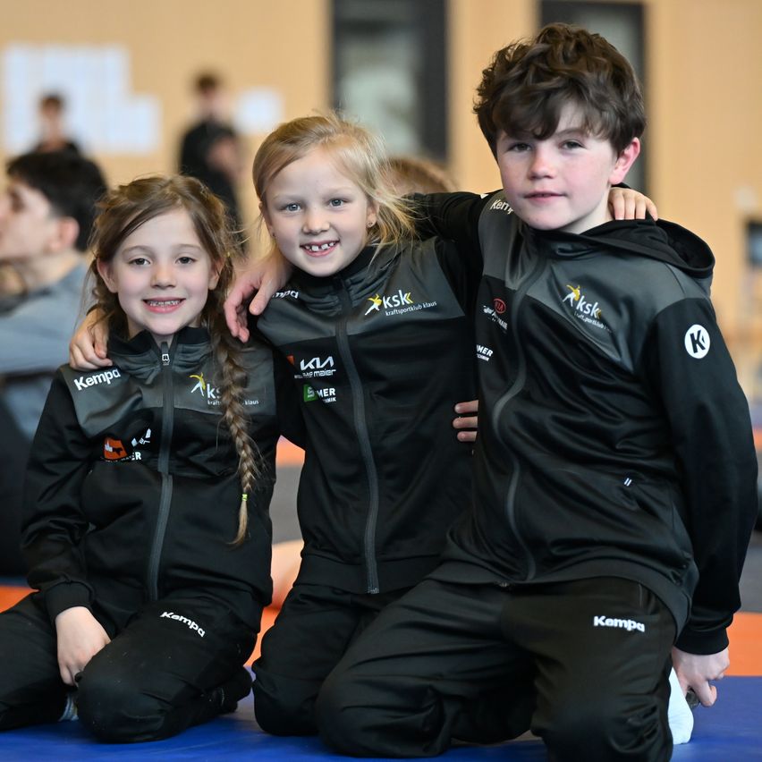 Three children in black sports uniforms sit together on a mat, smiling for a photo. Behind them, a group of people appears blurred, some standing and some sitting.