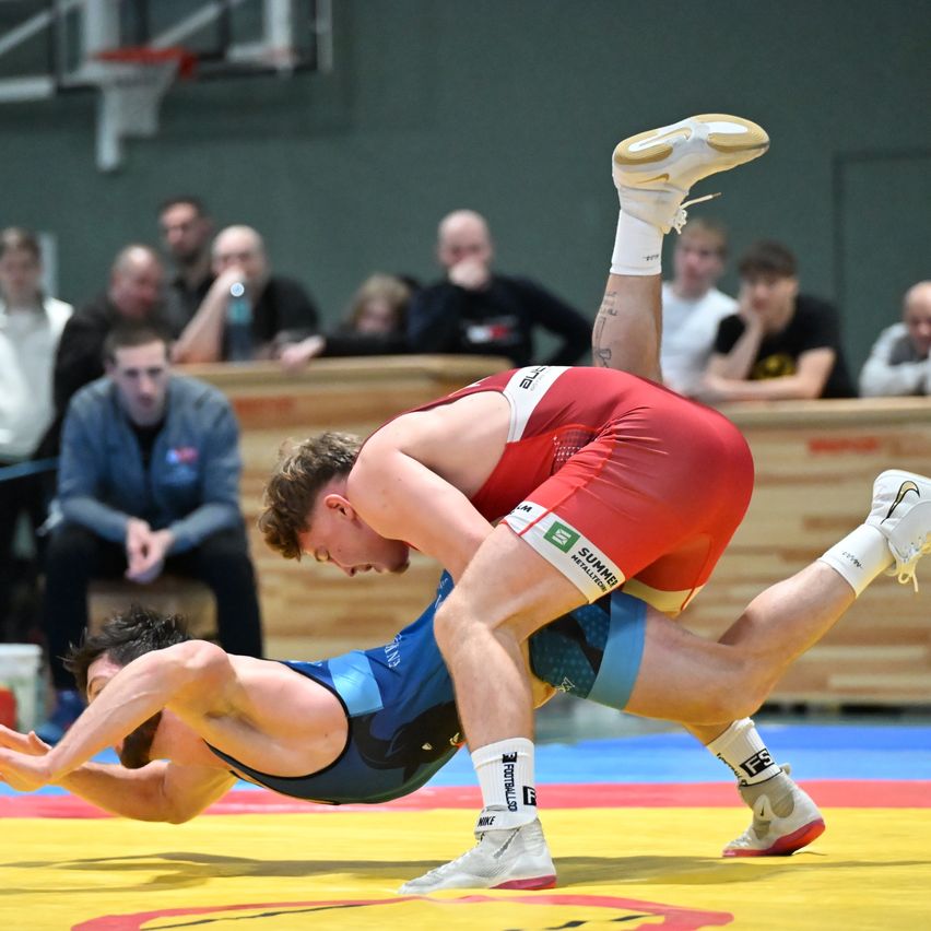 Two men wrestle on a mat in a gym; one wears red and the other blue. Spectators watch from benches.