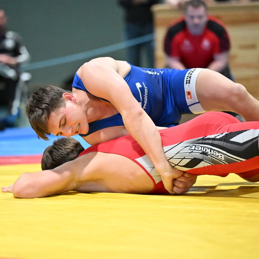 Two young wrestlers compete on a yellow mat. One in blue and white, the other in red and black. Spectators watch in the background.