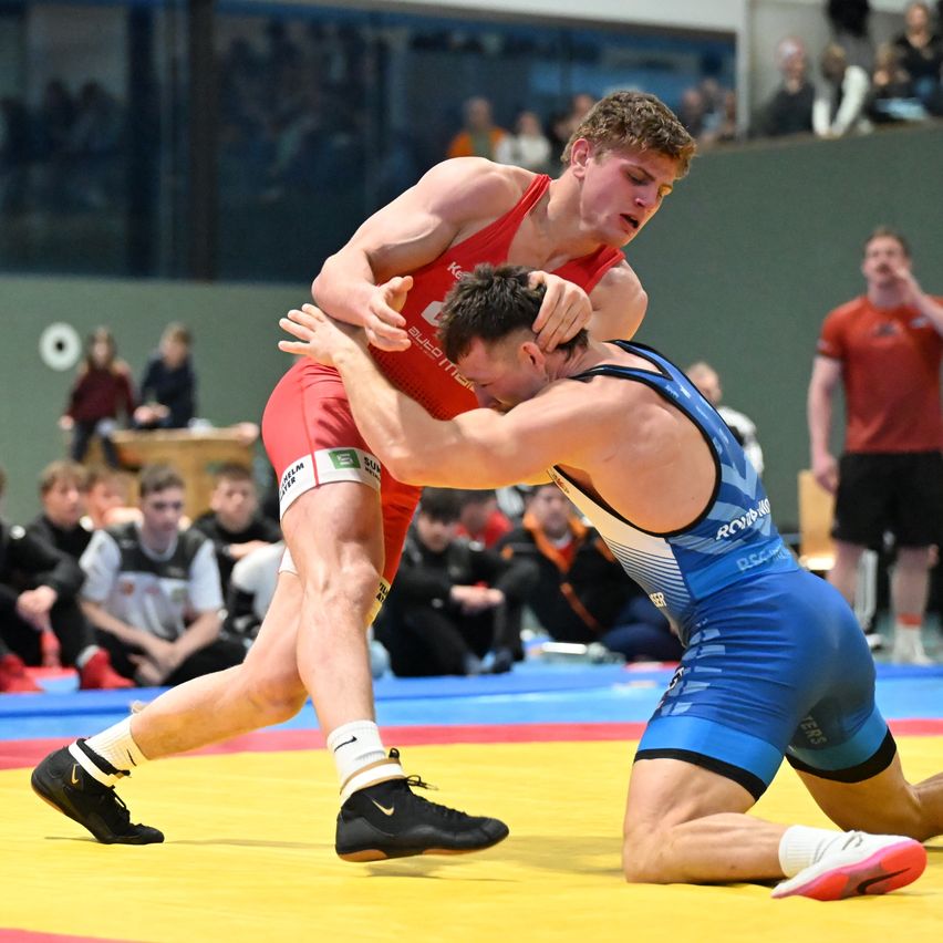 Two wrestlers in red and blue uniforms grapple in a wrestling match. The wrestler in red is on top. Spectators are watching from the bleachers.