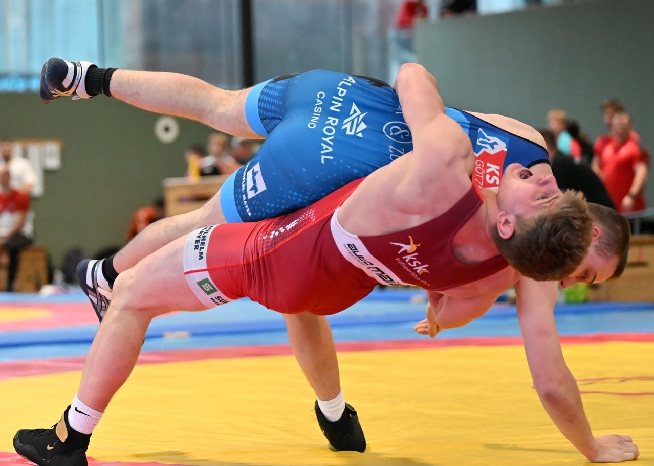 Two wrestlers in blue and red uniforms are in a wrestling match. The one in red has the upper hand, while the one in blue is defending. The match takes place on a yellow mat.