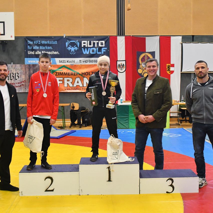 Three men and a woman stand on a podium with medals around their necks, with one holding a trophy. They are wearing sportswear, and behind them are flags and banners.