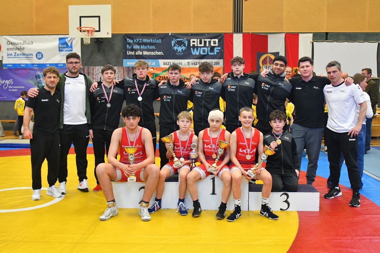 A group of young athletes with medals and trophies on a podium, posing for a photo. They are wearing red jerseys with numbers and medals. They are standing on a yellow mat.