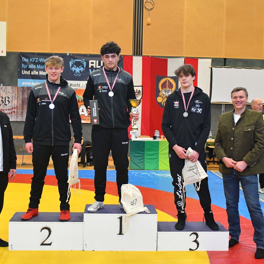 Three young men wearing medals stand on a podium. The man on the left has a number 2 on his podium, the middle one has a number 1, and the right one has a number 3. They are holding bags. A man is standing on the right.