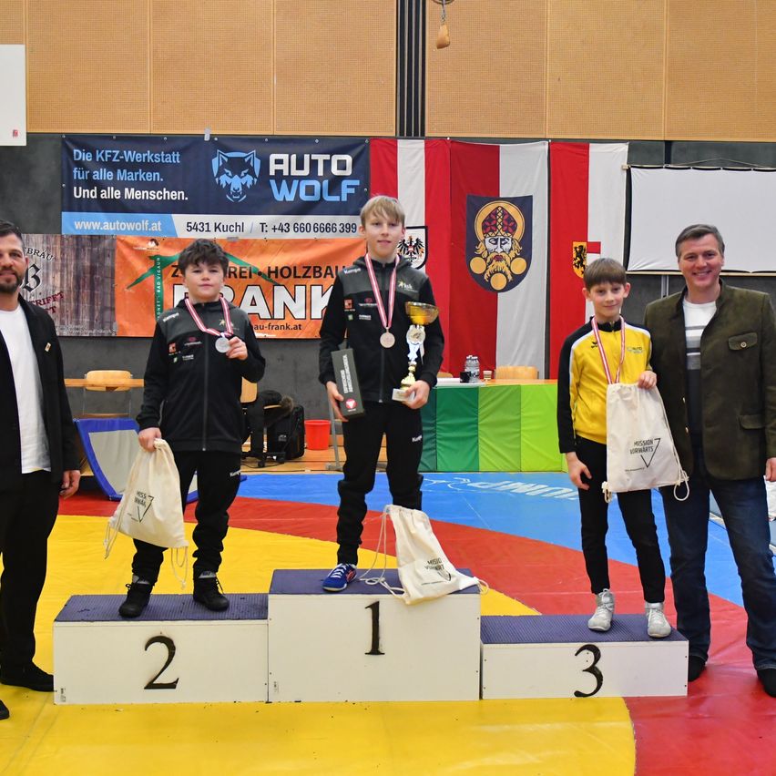 Three boys with medals stand on a podium with numbers 1, 2, and 3. A man stands beside them. The event is sponsored by AUTO WOLF.