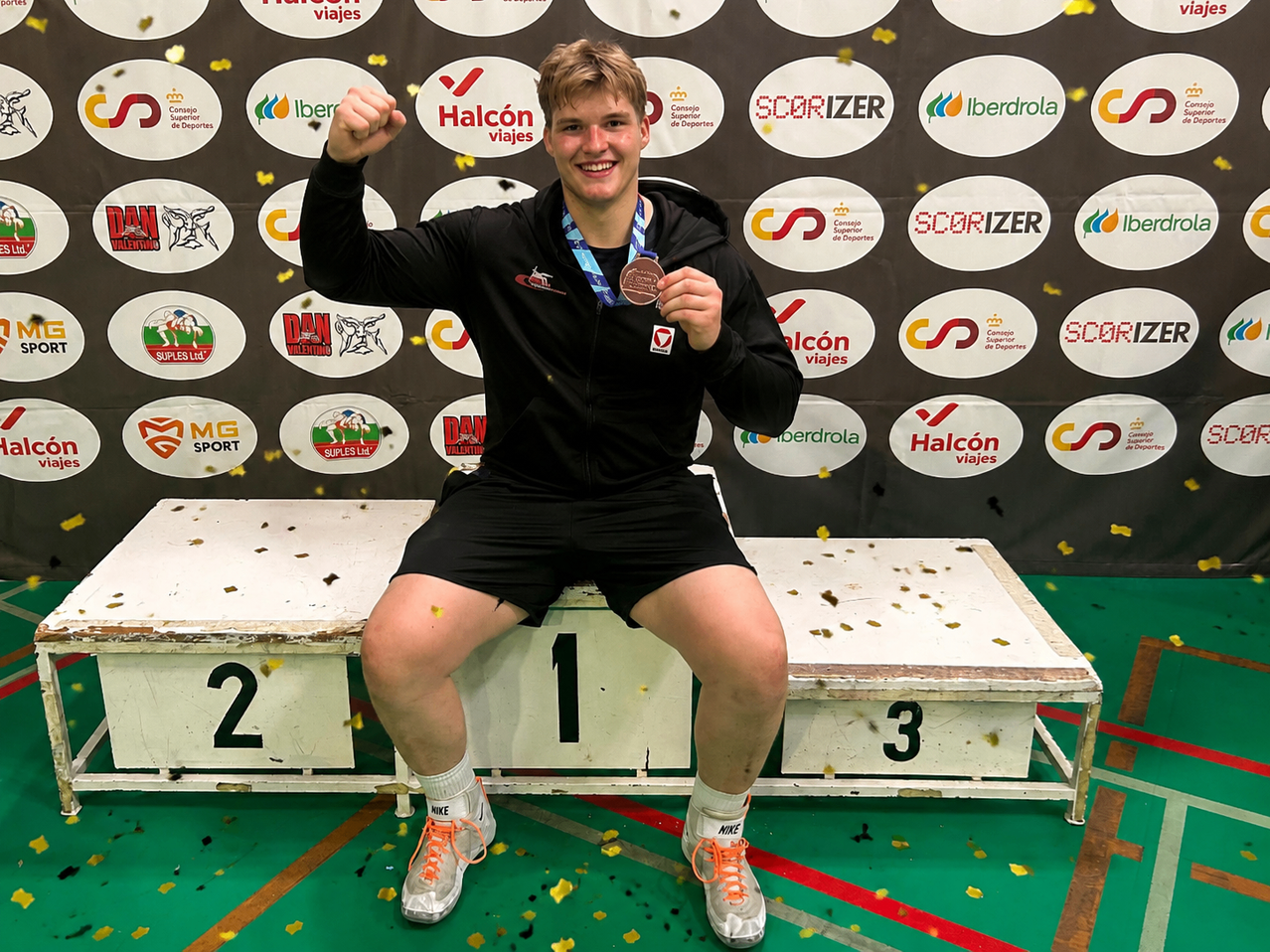 A man is sitting on a podium with a number 1 sign, wearing a black jacket, shorts, and sneakers. He is holding a medal in his right hand and raising his left hand. Behind him is a wall with logos and confetti.