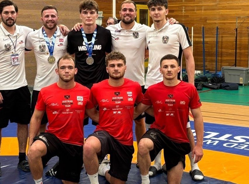 A group of male athletes wearing red and white uniforms pose for a photo on a wrestling mat. Some of them have medals around their necks.