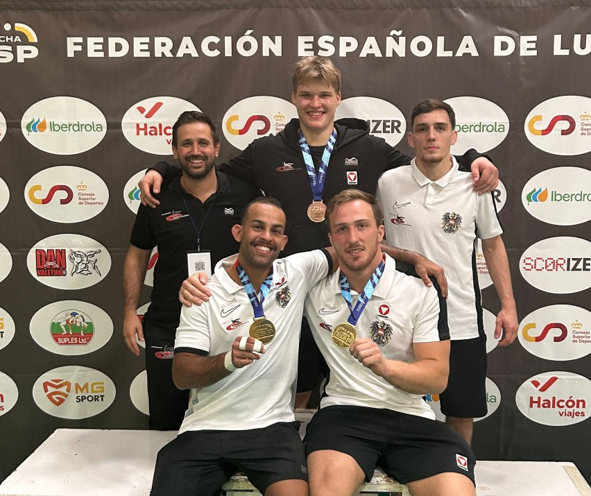 Five men pose for a photo with medals around their necks. Three of them are sitting on a bench, while two others stand behind them. They are all smiling and looking at the camera. Behind them is a wall with logos and text.