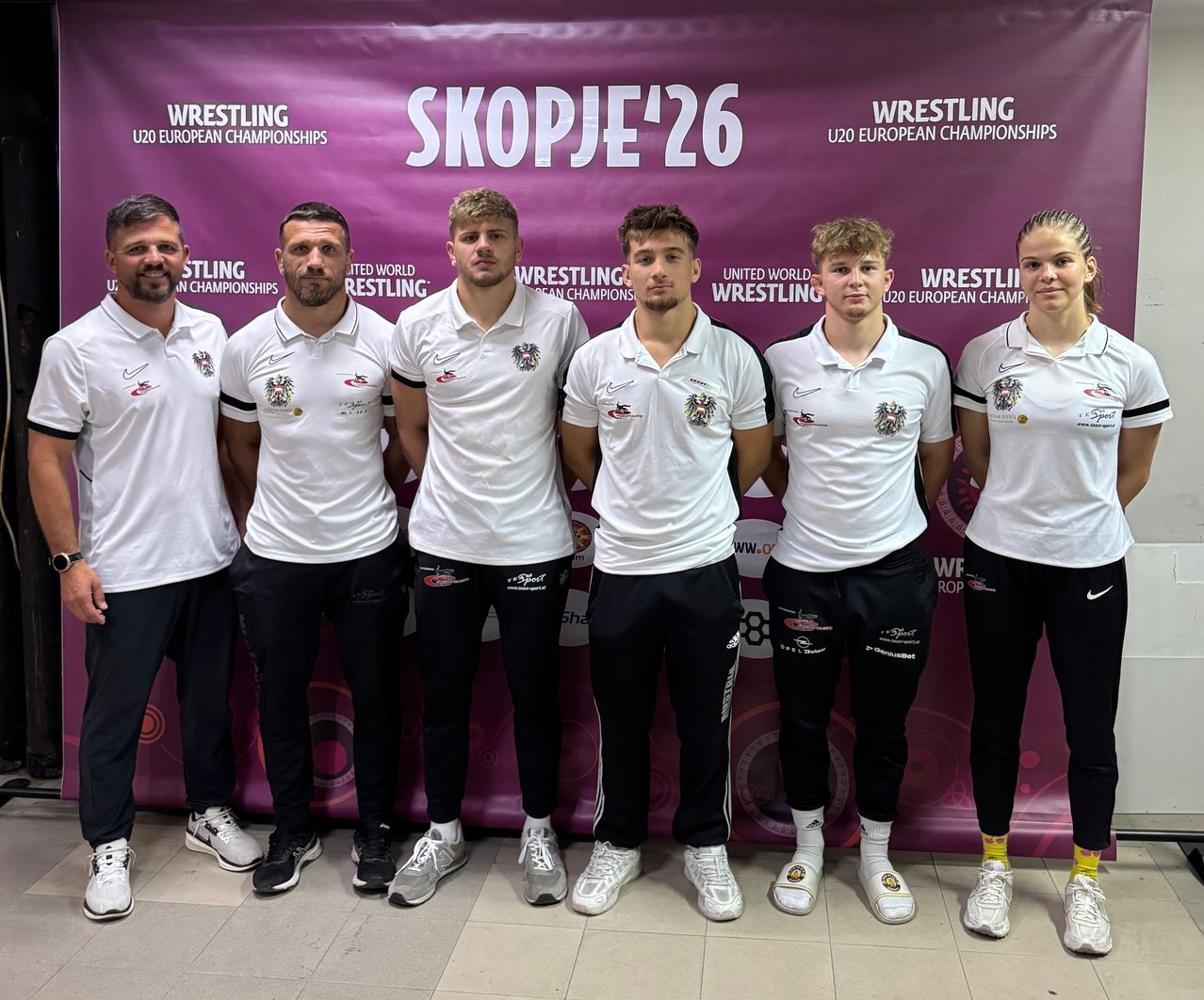 Six individuals dressed in white polo shirts with logos and black pants stand in front of a purple backdrop with the text 'Skopje'26' and 'Wrestling U20 European Championships'.