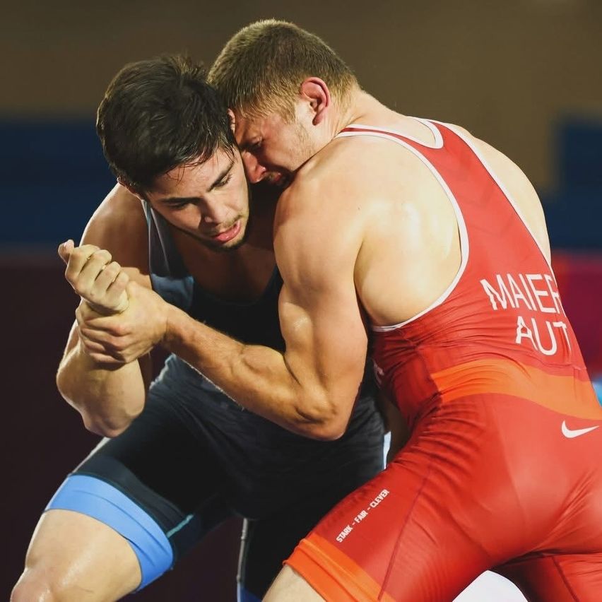 Two male wrestlers in a competition, one wearing a red singlet with 'MAIER' and 'AUT', the other in a blue singlet. They are in a grappling position.
