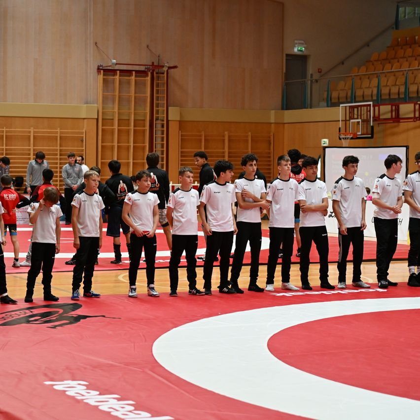 Eine Gruppe junger Jungen in weißen Shirts und schwarzen Hosen steht auf einem Wrestling-Matte in einem Sportsaal. Sie stehen der Zuschauertribüne gegenüber, während andere von den Seitenlinien zuschauen.