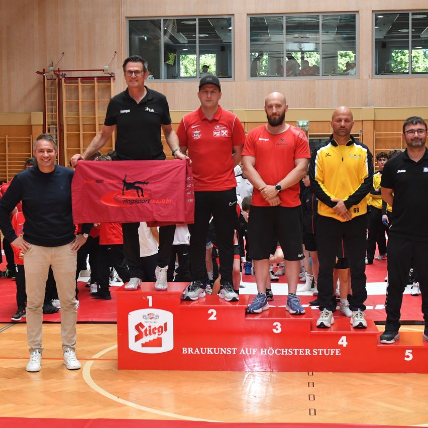 Fünf Männer stehen auf einem Podium in einer Turnhalle. Der Mann auf der linken Seite hält eine rote Fahne mit einem schwarzen Pferd. Das Podium hat die Zahlen 1 bis 5.