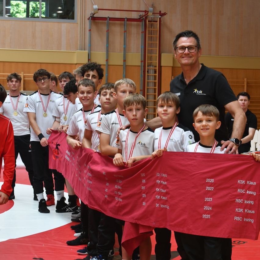 Eine Gruppe junger Jungen mit Medaillen um den Hals steht in einer Turnhalle, hält ein rotes Banner, mit einem lächelnden Mann in einem schwarzen Shirt hinter ihnen.