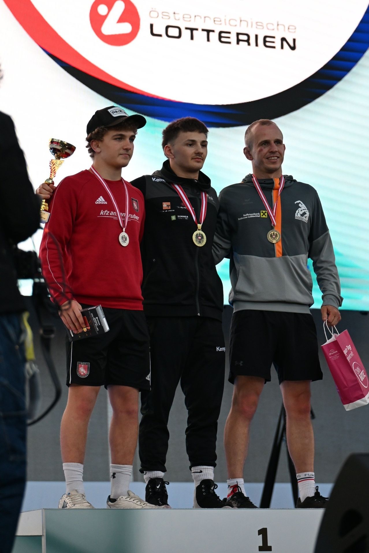Drei Männer auf einer Bühne, einer hält eine Trophäe, alle tragen Medaillen und lächeln. Einer hält ein Buch, der andere eine rosa Tasche. Dahinter ein verschwommener Hintergrund.