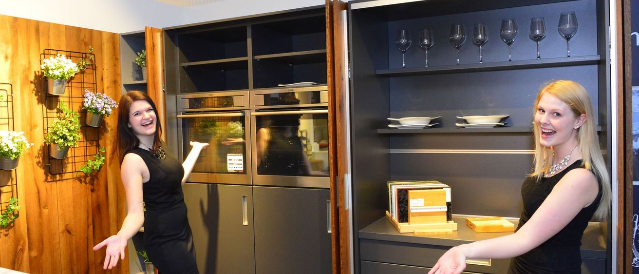 Zwei Frauen in schwarzen Kleidern stehen in einer modernen Küche mit grauen Schränken. Eine Frau zeigt auf einen Doppelofen, während die andere lächelt. Die Küche hat eine Holztür und Glasregale mit Weingläsern und Tellern.