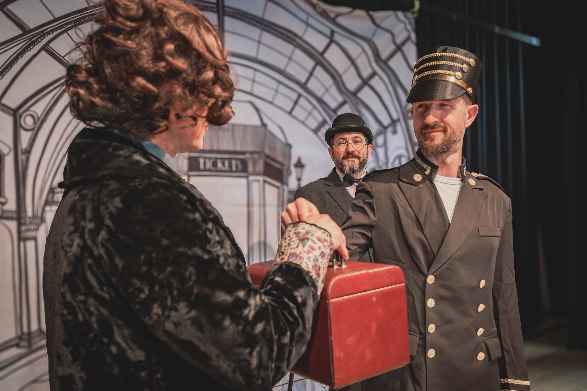 Zwei Männer in Uniform und eine Frau mit einer roten Aktentasche stehen auf einer Bühne und führen wahrscheinlich ein Theaterstück auf. Dahinter steht eine Wand mit der Aufschrift 'Tickets'.