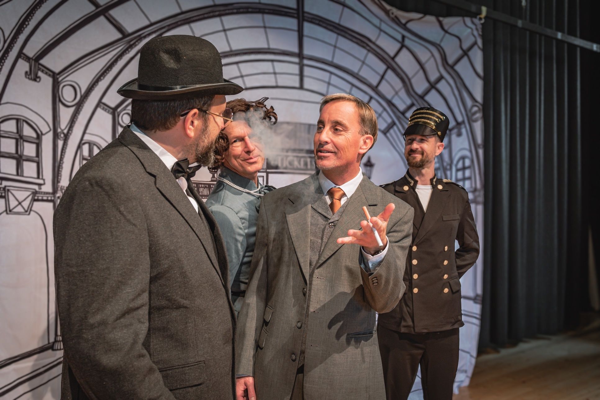 Vier Männer in Vintage-Kleidung stehen auf einer Bühne. Der Mann in der Mitte gestikuliert, während er spricht, und ein anderer raucht eine Zigarette. Der Mann im Hintergrund trägt einen Hut und eine Uniform.