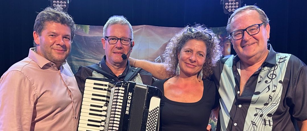 Vier Personen stehen lächelnd für ein Foto auf der Bühne. Ein Mann auf der linken Seite hält ein Akkordeon, und eine Frau in der Mitte ist barfuß. Ein Mann auf der rechten Seite hat ein Mikrofon. Hinter ihnen sind Stühle und ein Hintergrund mit Musiknoten.