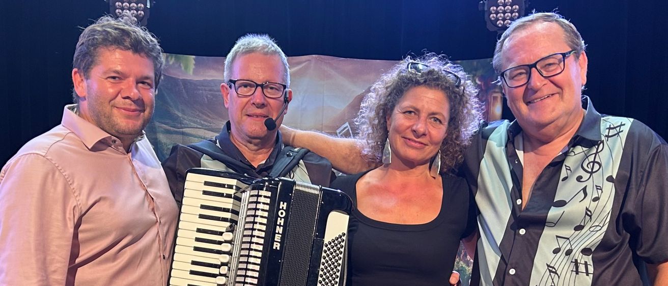 Vier Personen stehen lächelnd für ein Foto auf der Bühne. Ein Mann auf der linken Seite hält ein Akkordeon, und eine Frau in der Mitte ist barfuß. Ein Mann auf der rechten Seite hat ein Mikrofon. Hinter ihnen sind Stühle und ein Hintergrund mit Musiknoten.