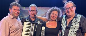 Vier Personen stehen lächelnd für ein Foto auf der Bühne. Ein Mann auf der linken Seite hält ein Akkordeon, und eine Frau in der Mitte ist barfuß. Ein Mann auf der rechten Seite hat ein Mikrofon. Hinter ihnen sind Stühle und ein Hintergrund mit Musiknoten.