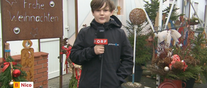 Ein junger Junge hält ein Mikrofon mit dem ORF-Logo in der Hand und steht vor einem Haus mit dekorativen Pflanzen und einem Schild mit der Aufschrift 'Weihnachten'.