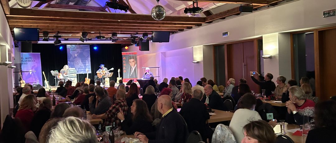 Ein Innenbereich mit Bühne, auf der Musiker auftreten. Das Publikum sitzt an Tischen, einige beobachten die Bühne, andere unterhalten sich. Der Raum ist mit rosa und blauen Lichtern beleuchtet.