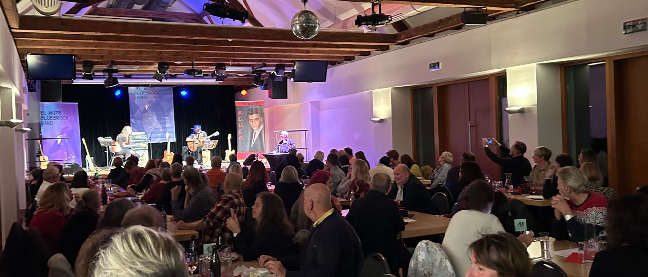 Ein Innenbereich mit Bühne, auf der Musiker auftreten. Das Publikum sitzt an Tischen, einige beobachten die Bühne, andere unterhalten sich. Der Raum ist mit rosa und blauen Lichtern beleuchtet.