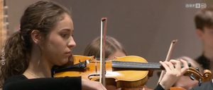Eine Frau in einem schwarzen Langarmkleid spielt Geige in einem Konzert, mit einem weiteren Geiger dahinter.