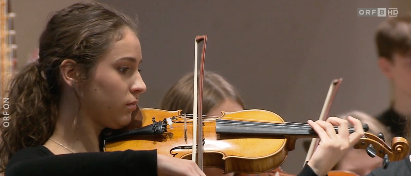 Eine Frau in einem schwarzen Langarmkleid spielt Geige in einem Konzert, mit einem weiteren Geiger dahinter.