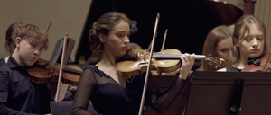 Eine Geigerin spielt in einem Konzert mit einem jungen Jungen, der eine Geige zu ihrer Linken spielt. Dahinter steht ein Flügel im Hintergrund.