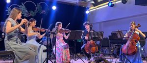 Fünf Musiker auf der Bühne, zwei mit Geige, einer mit Flöte und zwei mit Cello. Sie spielen mit Scheinwerferlicht auf ihnen.