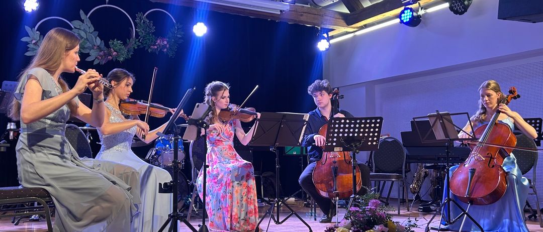 Fünf Musiker auf der Bühne, zwei mit Geige, einer mit Flöte und zwei mit Cello. Sie spielen mit Scheinwerferlicht auf ihnen.