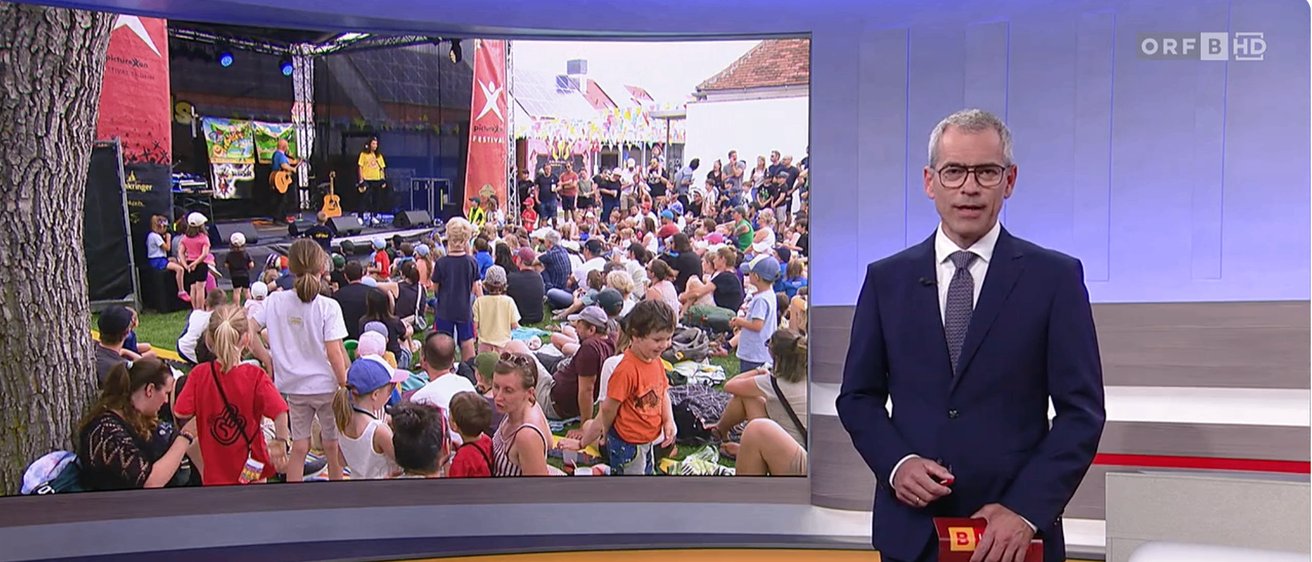 Ein Nachrichtensprecher berichtet von einem Festival und zeigt eine große Menschenmenge, die sich im Freien versammelt hat, einige sitzen und andere stehen. Im Hintergrund ist eine Bühne mit einem Künstler und einem Banner zu sehen.