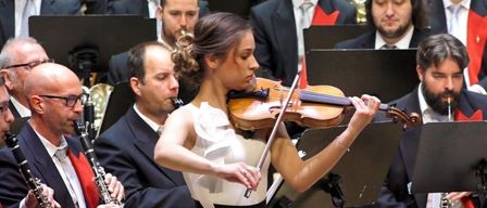 Eine Frau in einem weißen und schwarzen Kleid spielt Geige in einem Orchester. Hinter ihr spielen Musiker verschiedene Instrumente, darunter Klarinette und Posaune.