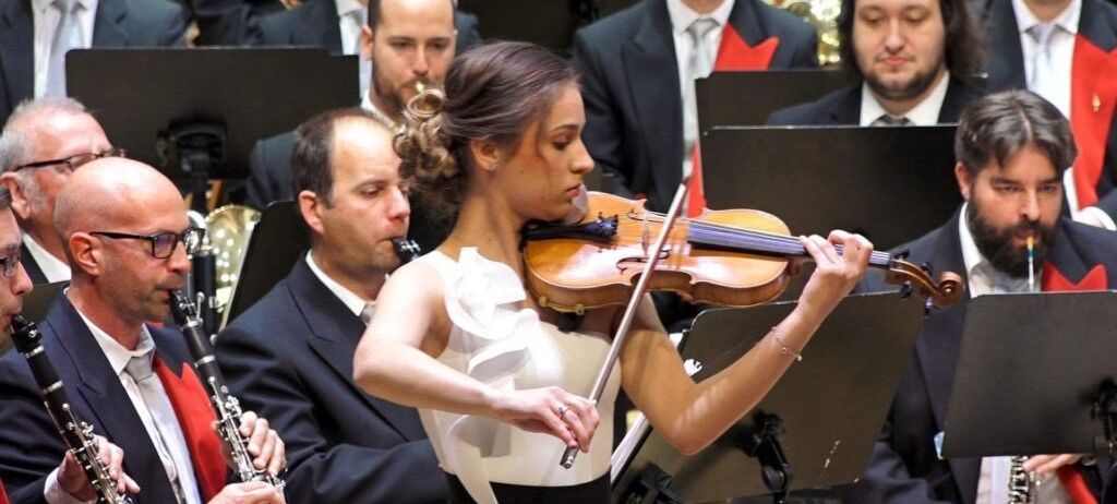 Eine Frau in einem weißen und schwarzen Kleid spielt Geige in einem Orchester. Hinter ihr spielen Musiker verschiedene Instrumente, darunter Klarinette und Posaune.