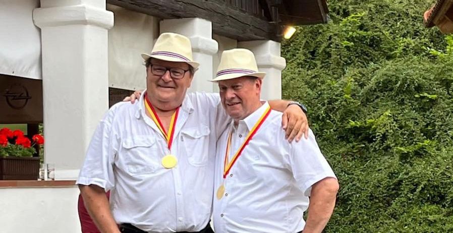 Zwei Männer in weißen Hüten und Medaillen posieren vor einem Gebäude mit einer Holzstruktur und Grünpflanzen im Hintergrund.