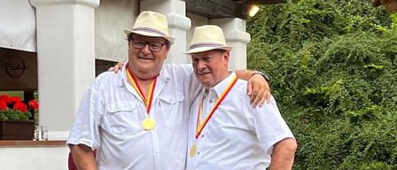 Zwei Männer in weißen Hemden und Hüten stehen eng beieinander und lächeln für ein Foto. Sie tragen Medaillen um den Hals. Ein Gebäude mit einem Holzdach steht hinter ihnen, und Grünpflanzen sind sichtbar.