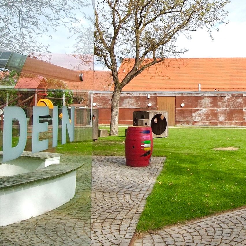 Ein Außenbereich mit einem gepflasterten Weg, der zu einem roten Fass mit bunten Designs führt, neben einem Baum und einem Gebäude mit orangefarbenen Dächern und Glasfenstern.